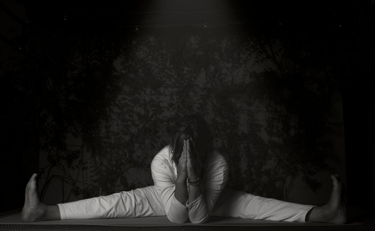 A woman practicing a yoga pose with a leg split indoors in monochrome lighting.